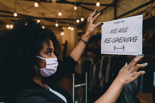 A woman wearing a cloth face mask attaches a sign to the front of a glass door. The sign reads "COME IN" in small letters above the larger declaration "WE ARE RE-OPENING." At the bottom of the sign is a diagram of two people standing at a distance from each other. In the background, the woman's shop is filled with racks of clothing and shoes.