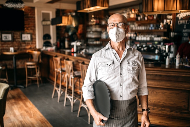  A man wearing a face mask stands in a restaurant and holds a serving platter. In the background is a well-stocked bar.