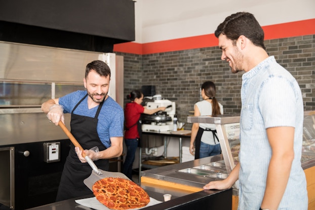 A pizzeria employee uses a metal pizza peel to place a pepperoni pizza on the restaurant's front counter. A customer on the other side of the counter watches with a smile. The employee is dark-haired with a beard, and he wears a black apron over a blue V-neck T-shirt. The customer is wearing a light-wash denim shirt. In the background, another employee operates an appliance in the kitchen while a third employee watches.