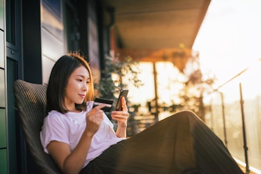 A young woman sits outside on a porch or balcony, reclining on a chair with her legs crossed. She holds her smartphone in one hand and a credit card in the other. In the background, the sky is lit gold by the sun.