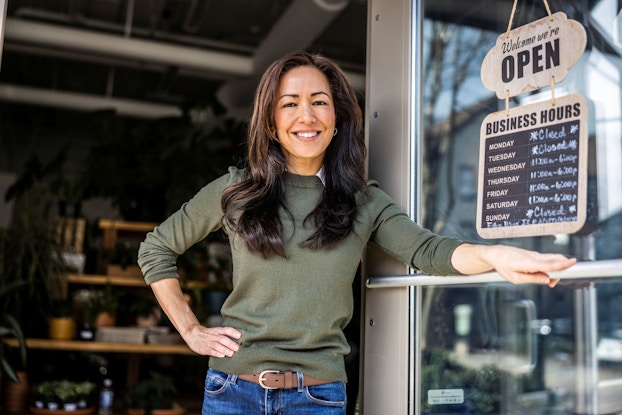 A female business owners wearing a green sweater stands proudly in front of her store. Next to her is an open sign listing her business's hours.