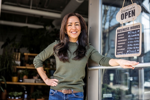  A female business owners wearing a green sweater stands proudly in front of her store. Next to her is an open sign listing her business's hours.