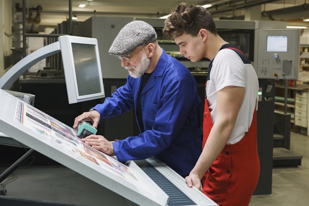  Two men, one older and white-bearded and one younger with dark curly hair, stand side-by-side at a drafting board. The older man, who wears a bright blue blazer and a gray newsboy cap, uses a handheld scanner to check the quality of the printout on the board. He holds the rectangular block-shaped scanner over one of the pictures on the printout. The young man, who wears red overalls over a white T-shirt, observes him.