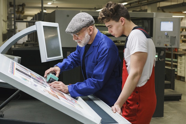 Two men, one older and white-bearded and one younger with dark curly hair, stand side-by-side at a drafting board. The older man, who wears a bright blue blazer and a gray newsboy cap, uses a handheld scanner to check the quality of the printout on the board. He holds the rectangular block-shaped scanner over one of the pictures on the printout. The young man, who wears red overalls over a white T-shirt, observes him.