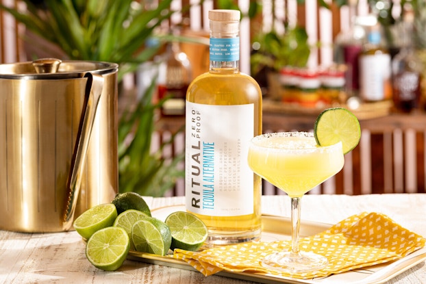 A bottle of Ritual Zero Proof's nonalcoholic tequila alternative, posed on a table next to a glass of the tequila (accented with a slice of lime on the rim), a brass ice bucket, and six halved limtes.
