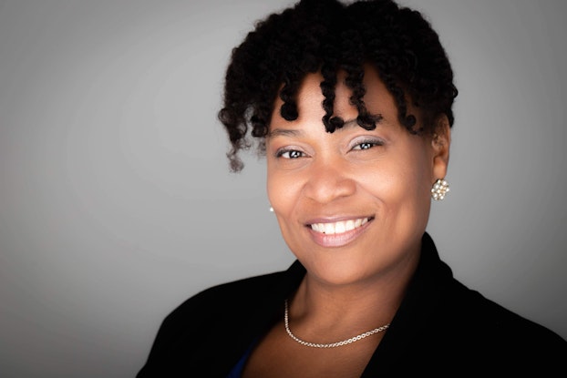 A head shot of a smiling Black woman. She has dark curly hair and dark brown eyes and she wears a black blazer and pearl cluster earrings.