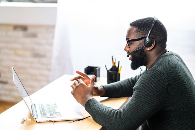 A man in profile sits at a desk facing an open laptop. He wears a headset and speaks into the attached microphone.