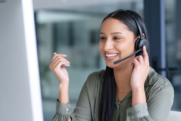 A head-and-shoulders shot of a young woman wearing a headset with an extended mouthpiece. She holds one finger to the earpiece of the headset and speaks into the mouthpiece with a smile.