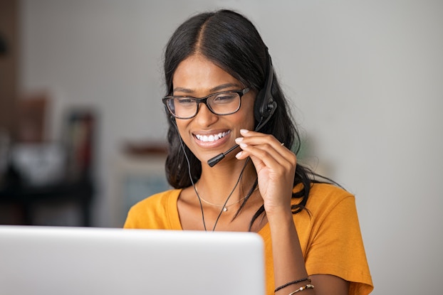 A woman wearing a headset sits at a laptop. One hand is on the mouthpiece of the headset to hold it near her mouth.