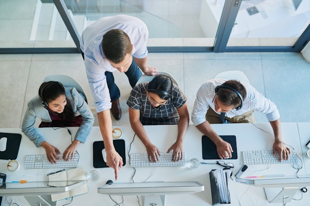  An overhead shot of a row of three sales employees wearing headphones. They are sitting in front of large computer monitors, into which the headphones are plugged. A keyboard and wireless mouse sits in front of each salesperson. A man in a white button-up shirt leans over the person in the middle and points at something on the computer screen.