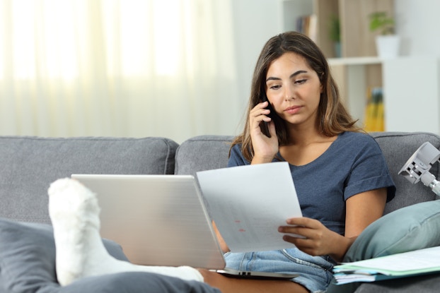A woma sits on a gray couch with a phone to her ear. She's looking at the piece of paper in her hand and her left leg is wrapped in a cast and elevated on a cushion in front of her.