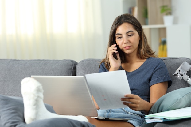  A woma sits on a gray couch with a phone to her ear. She's looking at the piece of paper in her hand and her left leg is wrapped in a cast and elevated on a cushion in front of her.