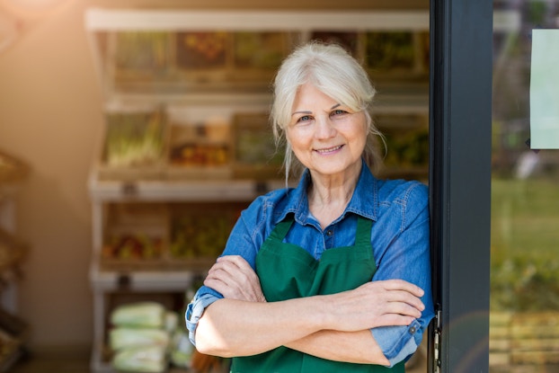 An older woman wearing a green apron stands in the doorway of a shop, smiling.