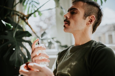 A close-up of a young man spraying a cologne or other fragrance on himself. The man has dark hair with shaved sides, a mustache, and a small goatee. His head is tilted slightly back as he sprays the fragrance mist towards his neck.