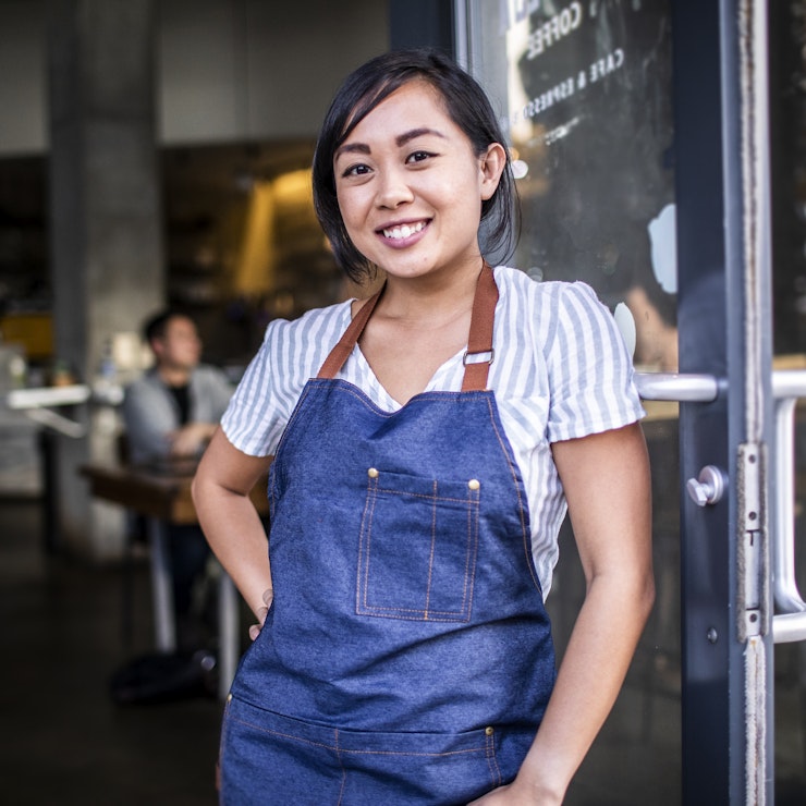 A young woman stands in the doorway of a restaurant or cafe, leaning against the glass-fronted door. She is wearing a white-and-light-blue-striped shirt under a denim apron. She has one hand on her hip and is smiling confidently.