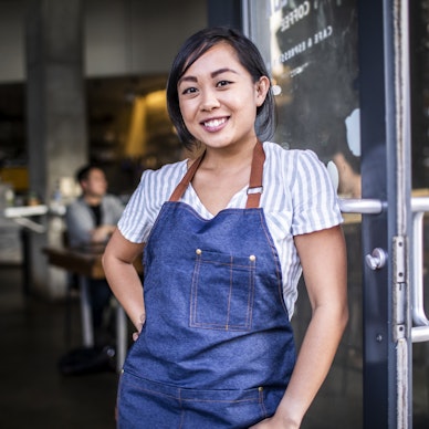 A young woman stands in the doorway of a restaurant or cafe, leaning against the glass door. She wears a white and light blue striped shirt under a denim apron. She has her one hand on her hip and her confident smile.