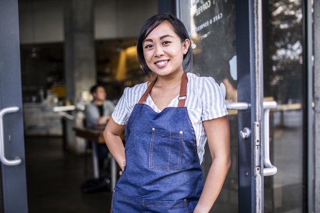  A young woman stands in the doorway of a restaurant or cafe, leaning against the glass-fronted door. She is wearing a white-and-light-blue-striped shirt under a denim apron. She has one hand on her hip and is smiling confidently.