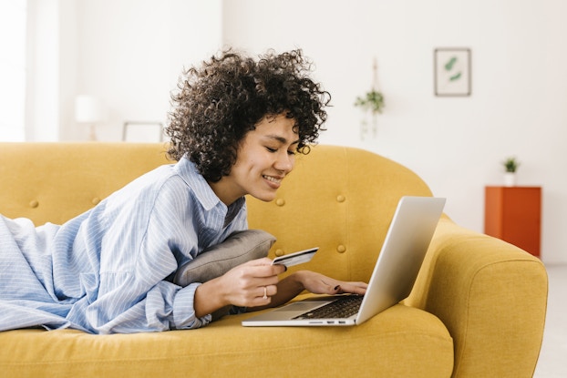  A curly-haired woman lies on her stomach on a faded yellow couch. She looks down at the open laptop in front of her on the couch with a smile. In one hand, she holds a credit card.