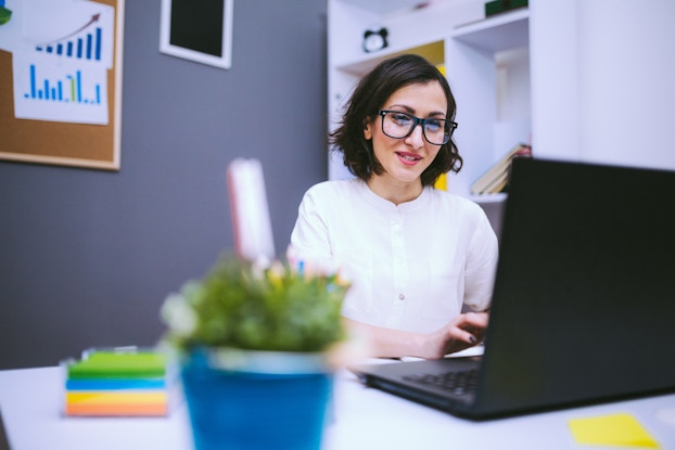 A woman wearing glasses sits at a desk and types on a laptop. The office around her is populated with a few charts on the walls and sticky notes and a potted plant on the desk.
