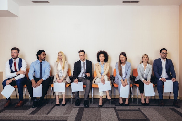  A row of people of many ethnicities and genders sit in chairs. They are all dressed in business casual wear and each person holds a piece of paper.