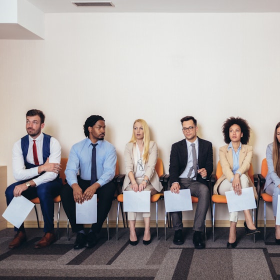  A row of people of many ethnicities and genders sit in chairs. They are all dressed in business casual wear and each person holds a piece of paper. 
