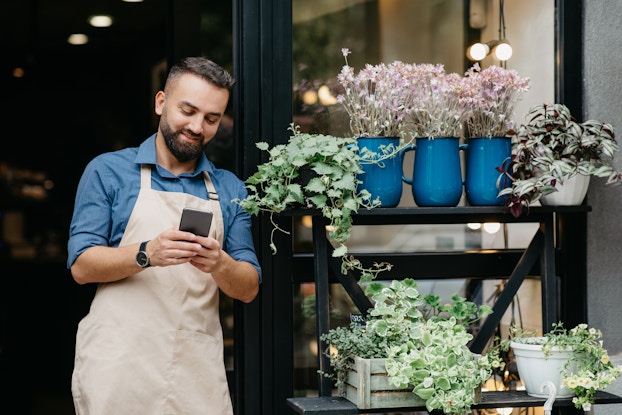  A man stands outside of a storefront and looks down at the smartphone in his hands. The man has dark hair and a dark beard, and he wears a khaki apron over a blue collared shirt with the sleeves rolled up. To the right of him is a two-tiered shelving set holding several potted plants.