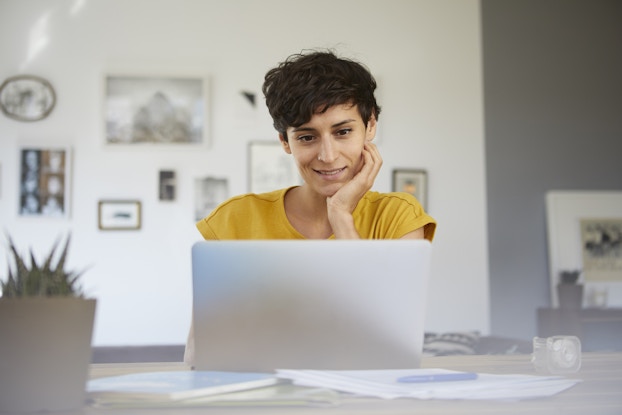  A woman with short dark hair sits at a table an open laptop. She has her head resting in one hand and a smile on her face. On the table around the laptop are some papers, a pen, and a notebook. A potted succulent plant sits on the table closer to the viewer, in the foreground.