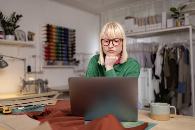  A woman sits at a table in front of an open laptop. She looks at the laptop screen with her chin resting on her fist. In the background is a white-walled room with a rack of clothes along one wall and rows of bobbins of thread in every color mounted on the other wall. The woman has shoulder-length white-blonde hair and wears a green shirt and red-rimmed glasses.