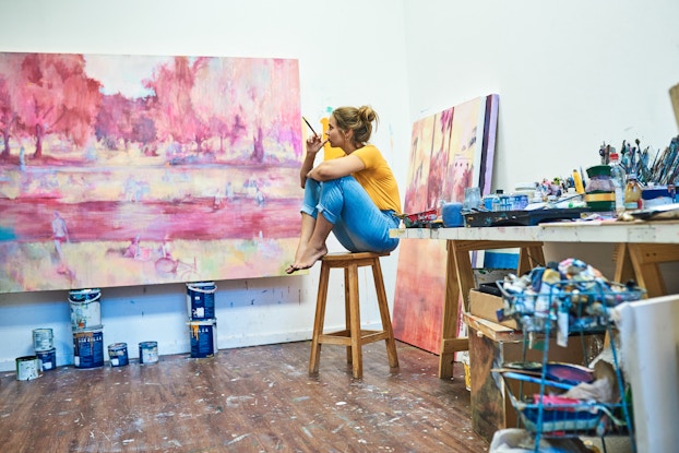  Woman artist sitting in her studio holding a paintbrush and looking at her painting.