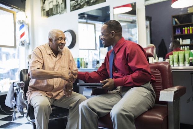  Two men, one older and one younger, sit in chairs in a barber shop and shake hands. The man on the left is the older one, with white hair and a white goatee. The man on the right is younger and wears a more formal outfit: a red button-up shirt, gray plants, and a blue tie. The younger man holds a digital tablet.