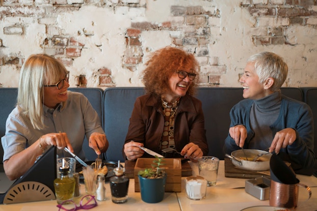 Three mature women seated side by side at a restaurant eat lunch and laugh.