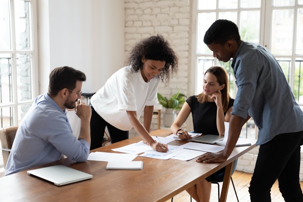  Four coworkers stand or sit around a table spread with various papers. One of the people standing, a woman in a white blouse, writes something on one of the papers as her colleagues look on.