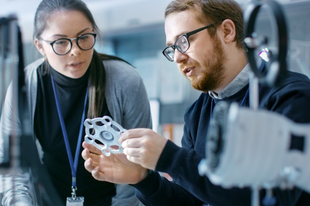 A bearded man wearing glasses holds up a 3D printed gear-like object while the woman standing next to him looks on. The object is flat and six-sided, and it somewhat resembles a snowflake. The man has his mouth open, as if he is explaining something. The woman is wearing glasses and a blue lanyard with an ID card at the end.