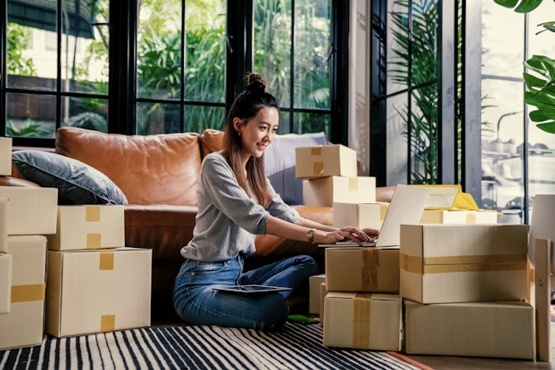 A woman sits cross-legged on a living room floor surrounded by taped-up cardboard boxes. She types something on a laptop perched on top of two small boxes. Behind her is a brown leather couch; behind the couch is a set of floor-to-ceiling windows, through which can be seen a yard filled with tall leafy ferns.