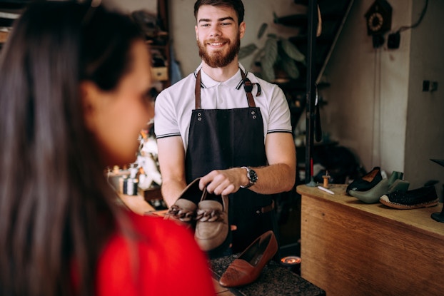 A young cobbler (shoemaker) holds out a pair of leather loafers to a customer in a shoe shop. The cobbler is a young man with brown hair and a brown beard; he wears a black apron over a white polo shirt. The shoes he presents are brown with a thick chain pattern across the tops. The customer stands out of focus in the foreground, with her back to the viewer; she has long brown hair and is wearing a red top. In the background, various shoes and boots sit on a wooden table in the shoe shop.