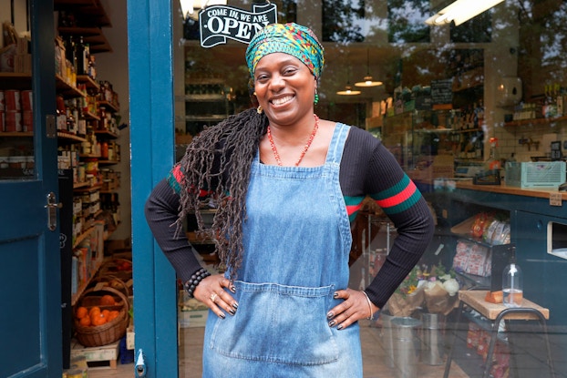  A Black woman entrepreneur stands outside her retail store and smiles at the viewer.