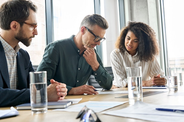 Three people in business casual wear sit at a conference room table and look down at a piece of paper at the table. The man sitting in the middle, who has graying hair and wears glasses, has one hand on his chin in deep thought. The woman on the right, who wears a beige-and-white striped shirt, appears to be speaking. The man on the left, who wears a checkered shirt and glasses, has his hands folded on the table. Also on the table are several glasses of water and a few other pieces of papers.