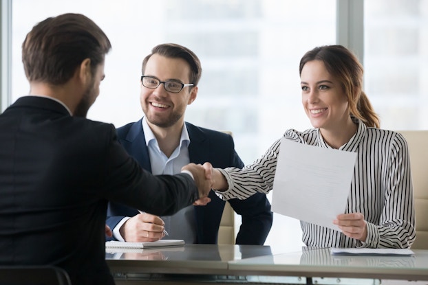  Three people in businesswear sit around a table. The two people on the side of the table facing the viewer are a man in glasses and a blazer and a woman wearing a black-and-white striped shirt. The woman holds a piece of paper and shakes hands with a second man sitting on the other side of the table.