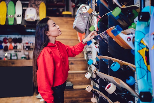 A young woman looks at a wall of skateboards in a skate shop. The boards are mounted on the wall at an angle, so their detailed undersides and colorful wheels are visible. The woman has long dark hair that reaches the middle of her back, and she wears a red hoodie and black pants. She reaches up with one hand to grab a skateboard at about eye level; the skateboard has blue transparent wheels and an orange and blue design on the underside.