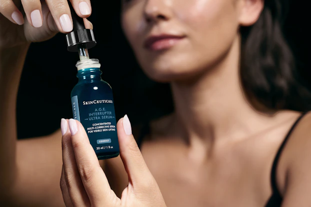 A woman holds a small blue vial of SkinCeuticals A.G.E. Interrupter Ultra Serum up to the camera while removing the eyedropper lid from the bottle.