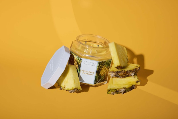 A Pineapple Passion candle from Slatkin + Co.'s ScentWorx line sitting with its lid off among several slices of pineapple. The candle is a golden yellow three-wick candle in an octagonal glass jar.