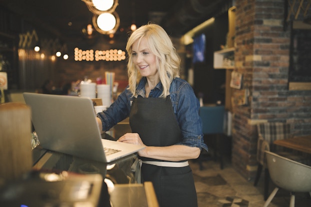  A woman stands at a counter in a restaurant and looks at the screen of an open laptop. The woman has long, wavy blonde hair, and she wears a black apron over a denim shirt. The restaurant in the background is out-of-focus and empty. Its walls are exposed brick and spherical lights hang from the ceiling.