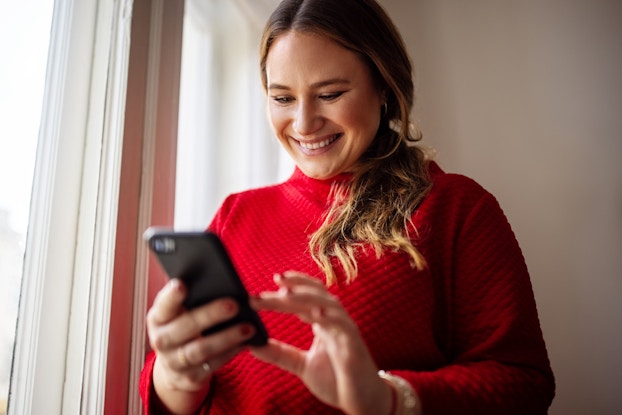  A smiling woman in a red sweater uses a smartphone. The woman has blonde hair that hangs over one shoulder.