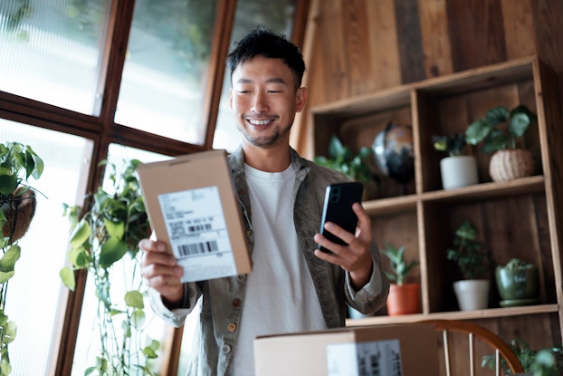  A young man holds a small package in one hand and a smartphone in the other. The package has a mail label on it, and the man looks at the box with a smile. In the background is a room filled with hanging and potted plants. One wall behind the man is made of floor-to-ceiling windows; the other wall is wood and lined with shelves that hold potted plants.