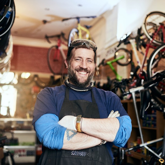 A bearded man stands in a storeroom, the walls of which are covered with handing and mounted bikes. The man wears a black apron over layered blue shirts and he has a tattoo of a triangle on one arm. The man has his arms crossed and smiles proudly.