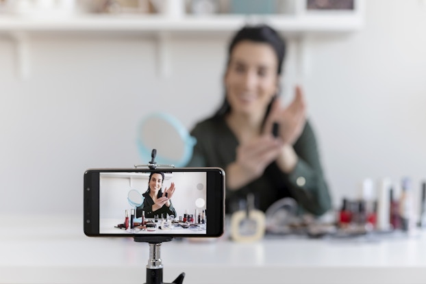A smartphone on a tripod takes a video of a woman showing off various beauty products.