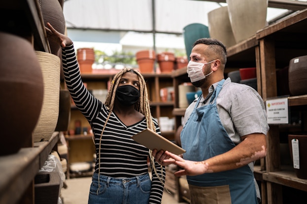 A Black woman gestures to a shelf of planters as she talks with an apron-wearing man with vitiligo. Both people are wearing face masks.