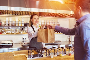  A woman in an apron stands behind a wooden counter and hands two paper bags to a customer. 