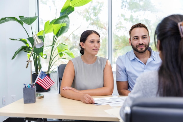 A man and a woman are seated in a bank office meeting with a loan manager to discuss a prospective business loan.