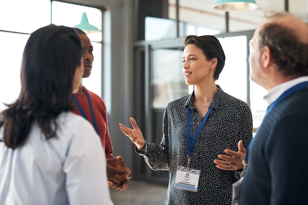  A group of four people of various ages, races, and genders stand in a loose circle in an open circle. All four are wearing blue lanyards with ID cards at the end. The person facing the camera is the focus of the group; they have short hair and they're speaking and gesturing as the other three people watch.